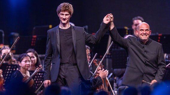 Jaap van Zweden (r), der Dirigenten des New York Philharmonic Orchestra, und Klaviersolist Jan Lisiecki (l) bedanken sich beim ersten Konzert im ehemaligen Kraftwerk Peenemünde auf der Ostseeinsel Usedom für den Applaus des Publikums. © picture alliance/dpa | Jens Büttner 