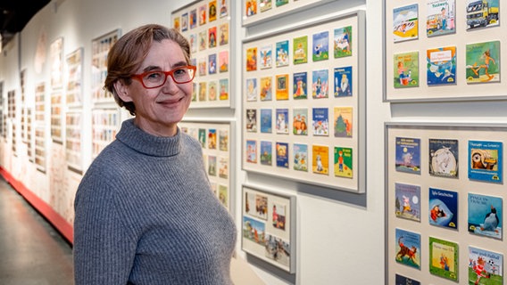 Eine Frau mit Brille und grauem Pullover steht lächelnd vor einer Ausstellung in Hamburg mit vielen Pixi-Büchern © Markus Scholz /dpa +++ dpa-Bildfunk Foto: Markus Scholz