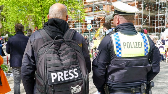 Ein Mann mit einem Rucksack, auf dem das Wort "Press" steht, wird von einem Polizisten begleitet. © IMAGO / BREUEL-BILD 