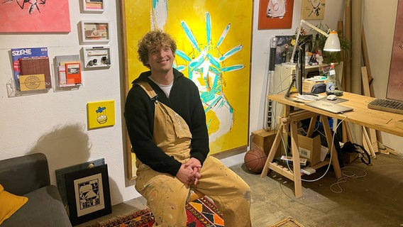 Der Künstler Mats Matthée sitzt auf einem Stuhl in seinem Atelier, hinter ihm hängen Bilder an der Wand. © NDR Foto: Anina Pommerenke