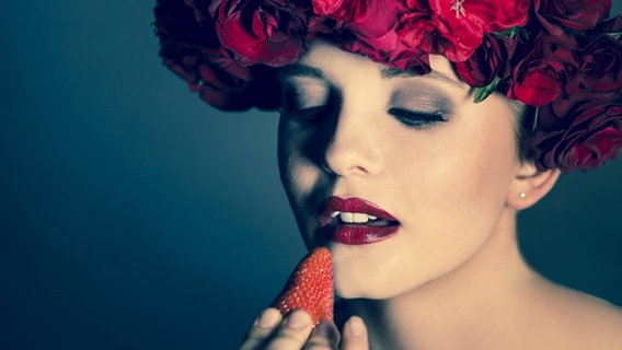 Eine Frau mit einem Blumenkranz im Haar und rot geschminkten Lippen isst eine Erdbeere. © imago Foto: Westend61