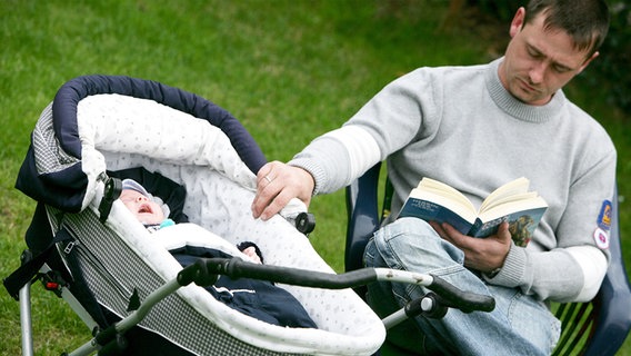 Vater sitzt mit einem Buch im Garten und betreut seinen sieben Wochen alten Sohn, der im Kinderwagen liegt. © dpa | Patrick Pleul Foto: Patrick Pleul