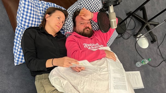 Ein Mann mit Vollbart und pinkem Hoodie und eine Frau in schwarzem Strickpullover liegen auf dem Fußboden unter einer Decke auf einem blau-weiß karierten Kopfkissen. Über ihnen hängt ein Mikrophon. © NDR Foto: Sarah Veith