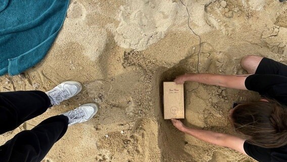 Im Sand stehen zwei Füße, eine Frau hockt sich nieder und scheint eine kleine Kiste einzugraben. Drum herum liegen zwei Handtücher und ein silberner Rucksack mit der Aufschrift "Air". © NDR Foto: Sarah Veith