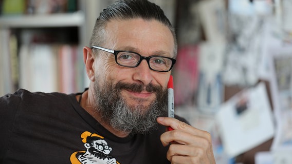 Der Comiczeichner Ralf König sitzt in seinem Atelier und hält einen roten Filzstift in der Hand. © picture alliance/Oliver Berg/dpa Foto: Oliver Berg
