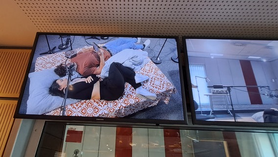 Zwei Personen liegen auf einer Matratze. Über ihnen hängt ein Mikrofon. Man sieht das Bild über einen Monitor. Es handelt sich um Studierende der Hochschule für Musik, Theater und Medien Hannover, die ein Hörspiel produzieren. © NDR 