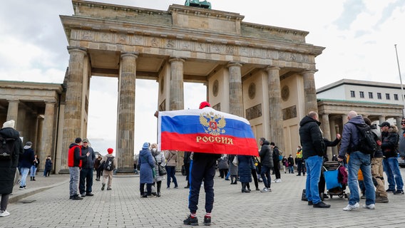 Ein Mann mit einer Flagge Russlands mit Wappen steht vor dem Brandenburger Tor, wo aus Russland stammende und in Deutschland lebende Menschen demonstrieren. © picture alliance/dpa | Gerald Matzka 