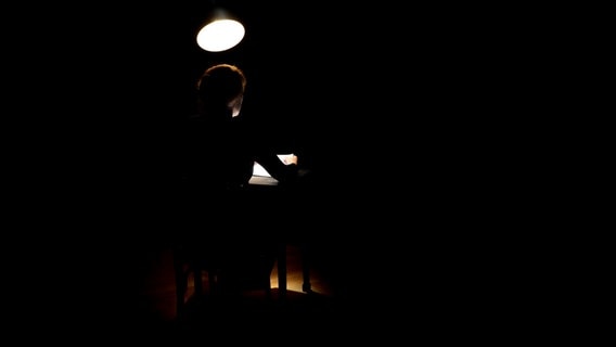 Martina Gedeck sitzt in einem komplett dunklen Raum an einem Tisch, der nur spärlich von einer Lampe beleuchtet wird. © Foto aus: "Samstagmittag, 12 Uhr", T. und P. Henke 