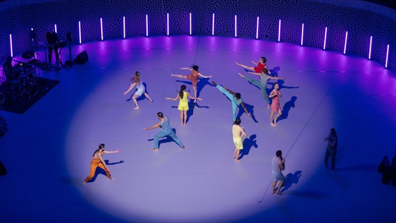 Bunt gekleidete Tänzerinnen und Tänzer bewegen sich auf einer blau-lila beleuchteten runden Bühne © Daniel Dittus Foto: Daniel Dittus