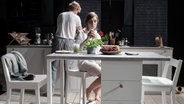 Ein Paar in einer Küche: Sie sitzt an einem Tisch und hält ein Glas in der Hand, er steht hinter ihr an einer Arbeitsplatte. © Armin Smailovic 