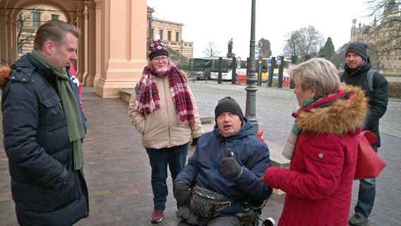 Eine Menschengruppe steht um einen Mann im Rollstuhl, der etwas erklärt. © NDR 