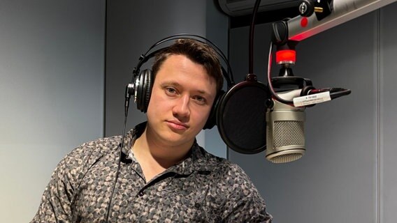Ein junger Mann sitzt mit Kopfhörern vor einem Mikrophon und blickt in die Kamera. Aufgenommen am 12.03.2025 im NDR Studio Göttingen © NDR 
