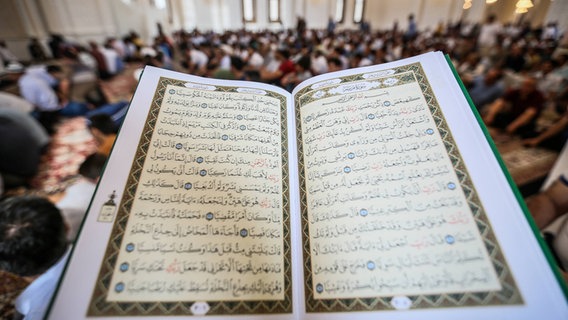 Zu sehen ist ein aufgeschlagener Koran , im Hintergrund sind Gläubige im Gebetsraum einer Moschee zu erkennen. © imago/ Pacific Press Agency 