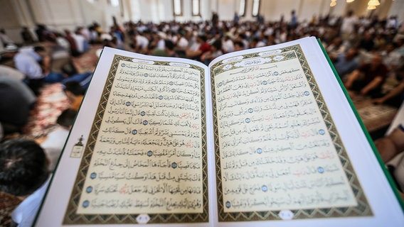 Zu sehen ist ein aufgeschlagener Koran , im Hintergrund sind Gläubige im Gebetsraum einer Moschee zu erkennen. © imago/ Pacific Press Agency 