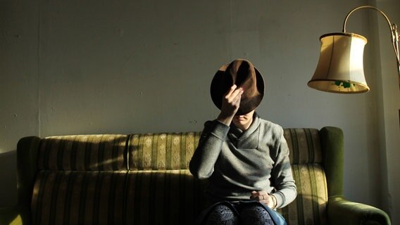 Eine Person sitzt auf einem altmodischen Sofa neben einer Stehlampe. Das Gesicht verdeckt sie mit einem Hut. © photocase / secretgarden Foto: secretgarden