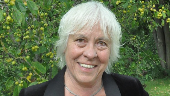 Eine Frau mit einer schwarzen Jacke und weißen Haaren lächelt in die Kamera. Im Hintergrund ist das Grün eines Gartens zu sehen © plan und rat 