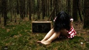 Eine Frau sitzt neben einem Koffer im Wald, das Gesicht von ihren Haaren verdeckt. © photocase / pixelhans Foto: pixelhans