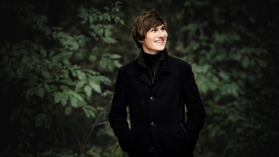 Ein junger Mann lächelt in die Ferne und steht vor grünen Büschen. Er trägt schwarze Klamotten. © Irène Zandel Foto: Irène Zandel