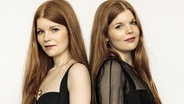 Die Zwillingsschwestern Clara und Marie Becker haben rote lange Haare und stehen mit dem Rücken zueinander, dabei schauen sie über die Seite nach vorne. © Clara & Marie Becker 