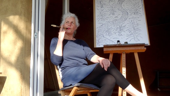 Miriam Wurster sitzt vor einer Leinwand mit einem Pinsel im Mund und schaut in die Ferne. © Miriam Wurster 