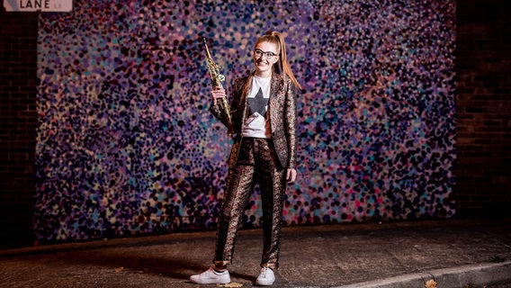 Jess Gillam steht mit ihrem Saxofone vor buntem Hintergrund © Robin Clewley Foto: Robin Clewley