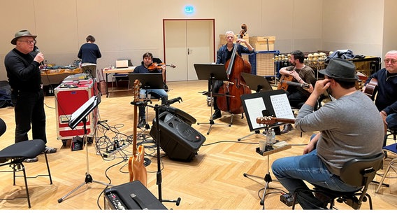 Links steht ein Mann und spricht in ein Mikrofon, rechts von ihm sitzen Musiker, die ihre Instrumente spielen. © NDR/Stefan Gerdes 