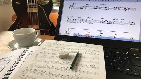 Auf einem Schreibtisch sind Noten auf Papier und auf dem Bildschirm eines Laptops zu sehen. Im Hintergrund steht eine Gitarre. © Jo Kappl Foto: Jo Kappl