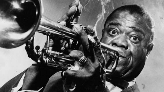 Jazz Trompeter und Sänger Louis Armstrong, spielt seine Trompete mit Zigarette in der Hand und schaut mit typisch aufgerissenen Augen in die Kamera. Jazz Trompeter und Sänger Louis Armstrong, spielt seine Trompete mit Zigarette in der Hand und schaut mit typisch aufgerissenen Augen in die Kamera. © picture alliance / ZUMAPRESS.com | Foto: Bob Moreland