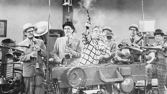 Der amerikanische Bandleader Spike Jones schießt mit einem Colt in die Luft und gibt den charakteristischen Startschuss für seine Band City Slickers, schwarz-weiß Aufnahme von 1954. © picture alliance / Everett Collection 