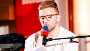 Ein jüngerer Mann mit Brille sitzt vor einem Mikrofon und liest aus einem Buch. © Literarischer Salon Hannover / Nerea Lakuntza Foto: Nerea Lakuntza