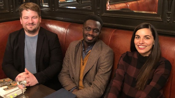 Alexander Solloch, Mohamed Mbougar Sarr und Franziska Herrmann sitzen an einem Tisch und blicken fröhlich in die Kamera. © Hanser Verlag Foto: Henriette Kuch