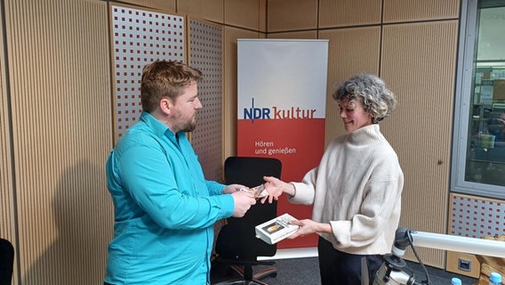 Alexander Solloch gibt Lisa Kreißler Geld für ein Buch, welches Lisa Kreißler in der Hand hält. © NDR 