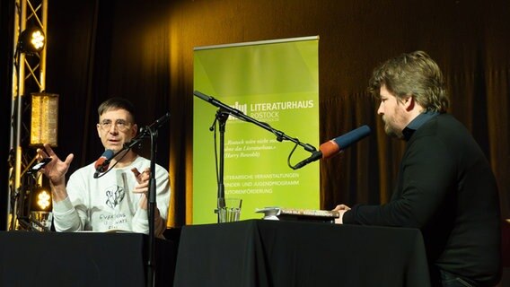 Tijan Sila und Alexander Solloch sitzen an Tischen auf einer Bühne. © Reiner Mnich Foto: Reiner Mnich