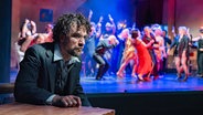 Ein niedergeschlagen aussehender Mann sitzt an einem Tisch, während im Hintergrund Menschen ausgelassen tanzen. © Thomas Ulrich 