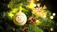 An einem Weihnachtsbaum hängt eine Kugel mit wütendem Smiley. © picture alliance / CHROMORANGE | Christian Ohde 