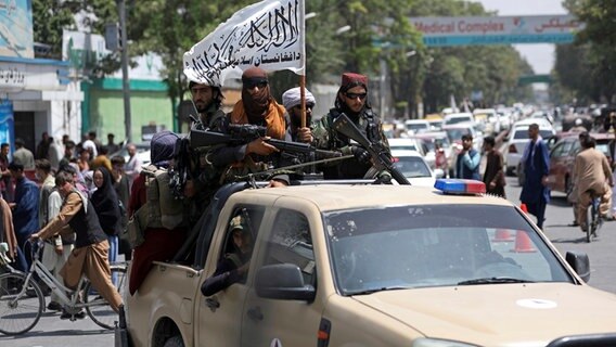 Schwer bewaffnete Taliban-Kämpfer patrouillieren nach ihrer Machtübernahme mit wehender Fahne des Islamischen Emirats Afghanistan durch Kabul. © picture alliance/dpa/AP Foto: Rahmat Gul