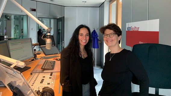 Zwei Frauen mit dunklem Haar und breiten Lachen stehen neben einem Schreibtisch über dem ein Mikrofon hängt. © NDR Foto: Stephan Sturm