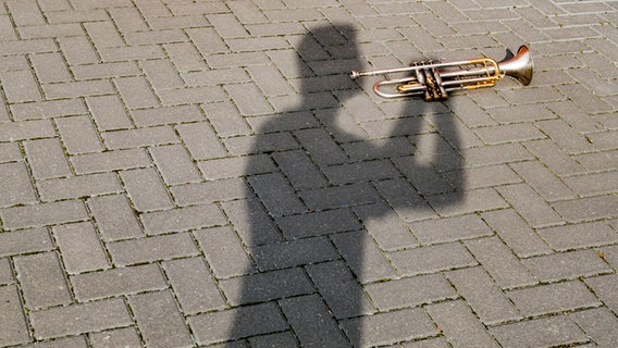EIne Trompete liegt auf dem Boden auf dem Schatten eines Trompetenspielers. © photocase Foto: David-W