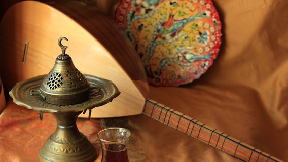 Eine Baglama, ein Glas Schwarztee und eine orientalische Lampe stehen auf einer goldbestickten Decke. © IMAGO / Pond5 Images 