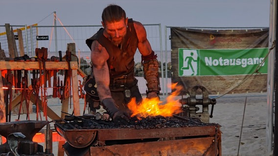 Ein Mann in Lederrüstung beugt sich im Freien über ein Feuer. © NDR Foto: Peter Schanz