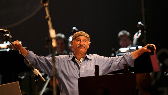 Jazz-Pianist Joe Zawinul steht lächelnd auf der Bühne zwischen Mikrofonen und Notenständern. © imago/Skata 