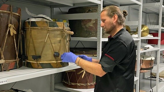 Ein Mann, der Gummihandschuhe trägt, steht vor einem Regal, in dem alte Trommeln lagern. Ein Mann, der Gummihandschuhe trägt, steht vor einem Regal, in dem alte Trommeln lagern. © NDR Foto: Lydia Callies