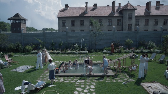 Eine große Familie springt auf der grünen Wiese um einen kleinen Pool herum - dahinter ist ein KZ zu erkennen. Szene aus dem britisch-polnischen Spielfilm "The Zone of Interest" von Jonathan Glazer © Leonine 