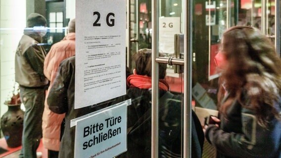 Besucher einer Vorstellung der Hamburger Kammerspiele gehen am Eingang an einem Aushang vorbei, der auf die geltenden 2G-Regeln im Haus für den Theaterbesuch hinweist. © picture alliance/dpa | Markus Scholz 