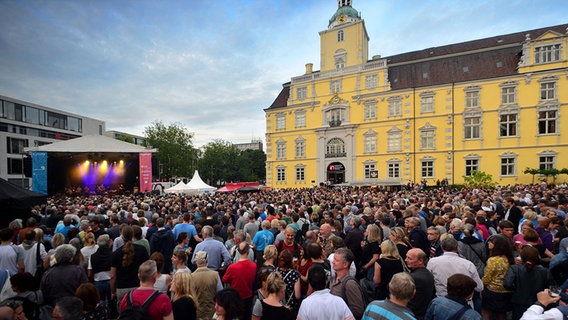 Ein gelbes Schloss im Hintergrund, davor stehen hunderte Menschen und schauen auf eine Bühne, die links im Bild steht. © Kultursommer Oldenburg 