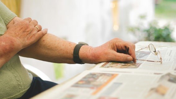 Der Arm einer älteren Frau ruht auf einem Tisch, daneben liegen eine aufgeschlagene Zeitung und eine Lesebrille. © Photocase Foto: poly picture