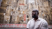 Der ghanaische Künstler Ibrahim Mahama steht in Osnabrück vor einer von ihm mit Jutesäcken verhüllten Fassade. © picture alliance / epd-bild Foto: Detlef Heese