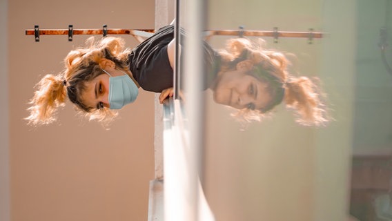 Eine junge Frau lehnt sich aus einem Fenster.  Sie trägt eine Mund-Nasen-Bedeckung.  In der Spiegelung des Fensters sieht man sie ohne sie lächeln.  © photocase.de Foto: Andoni