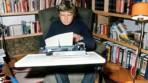 Hardy Krüger sitzt in seinem Sessel an einem Tisch mit einer Schreibmaschine und schreibt an einem Roman. © dpa / picture alliance Foto: Klar