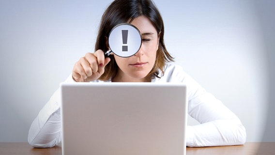 Eine Frau Blickt mit einer Lupe, auf der ein Ausrufezeichen zu sehen ist in einen Laptop. © iStock Foto: muharrem öner